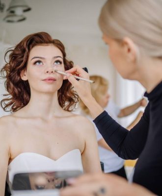Valmiera, Latvia - September 9, 2023 - Bride getting her makeup done by a makeup artist, preparing for the wedding, with her wedding gown hanging in the background.